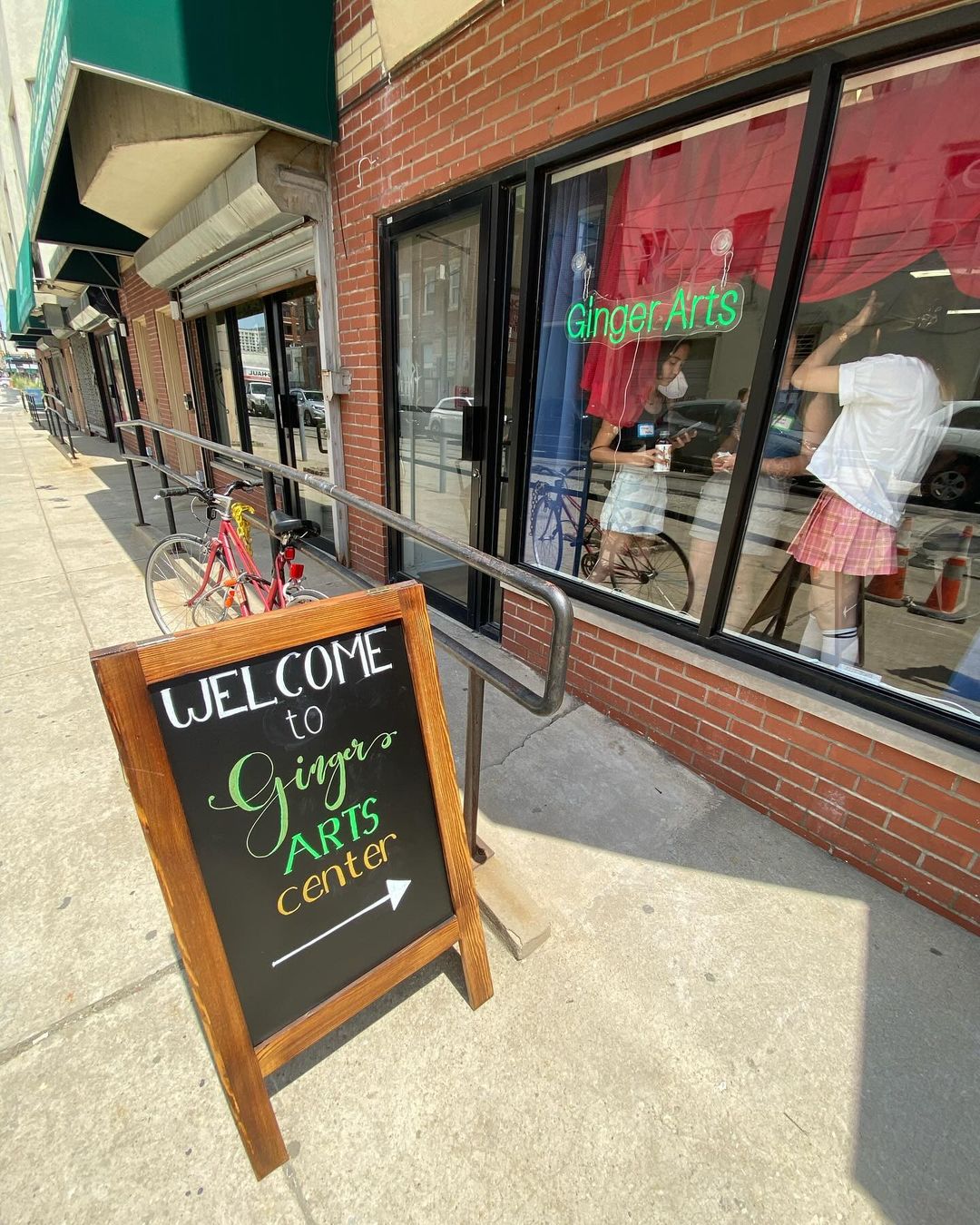 A sandwich board on the sidewalk reads "Welcome to Ginger Arts Center" with an arrow pointing towards the red brick entrance. A neon "Ginger Arts" sign glows in the window and young people are gathered inside.